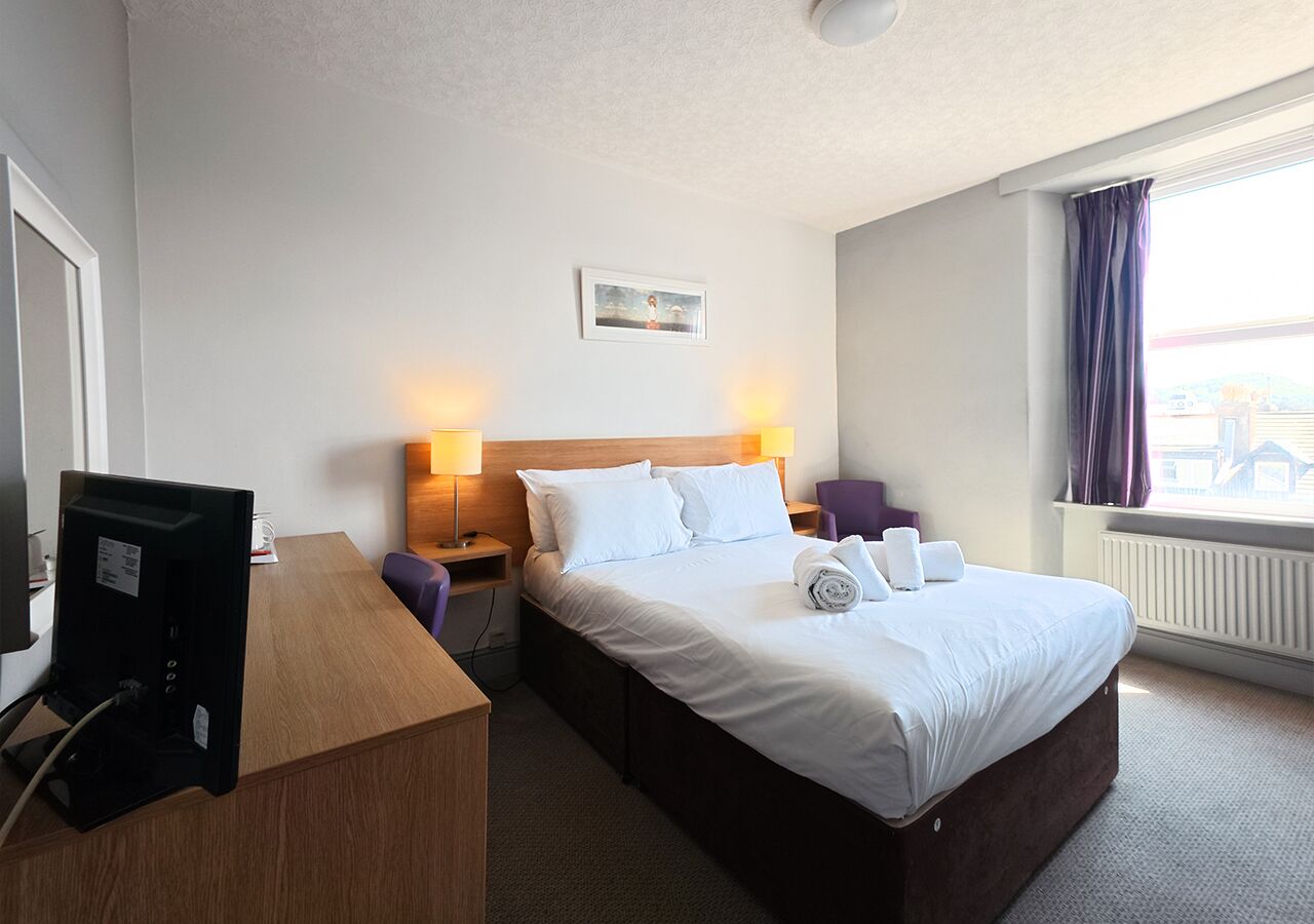 Una habitación de hotel con cama, lámparas de noche, escritorio con ordenador y ventana con cortinas.