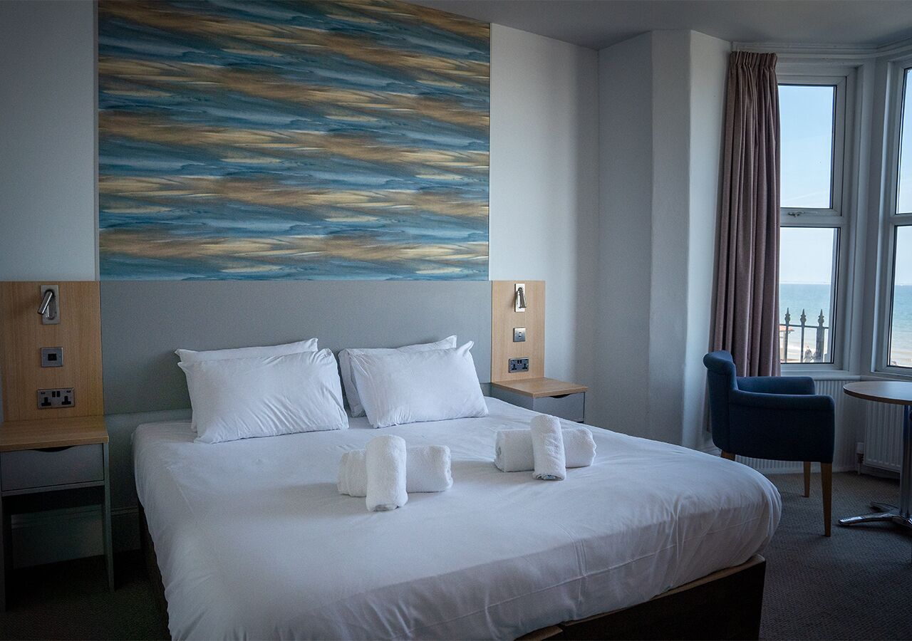 A hotel room with a large bed, two bedside tables, a chair, a window with curtains, and a wall with a blue abstract painting.