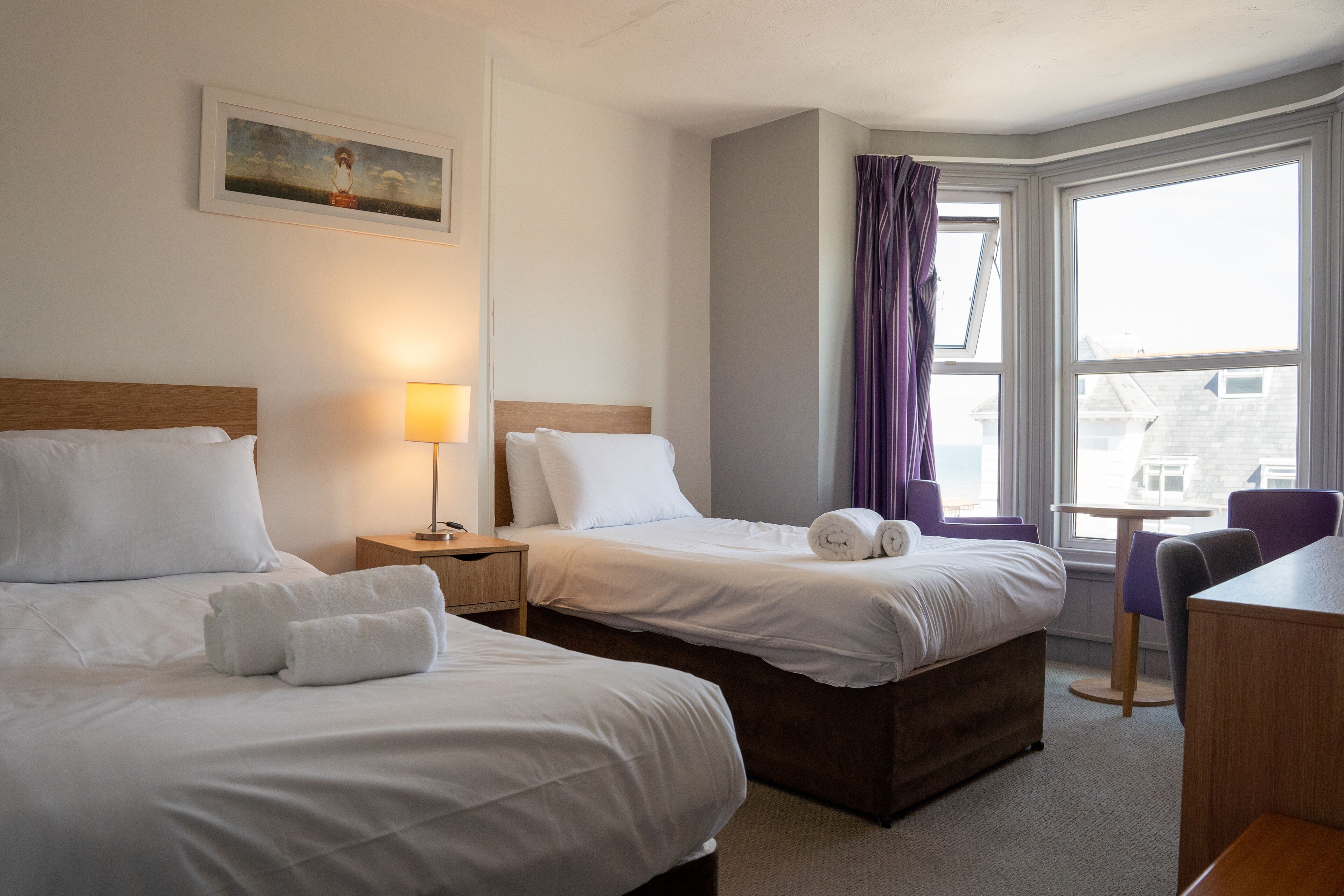 Habitación de hotel con dos camas, un escritorio, una silla, una ventana con cortinas y un cuadro en la pared.