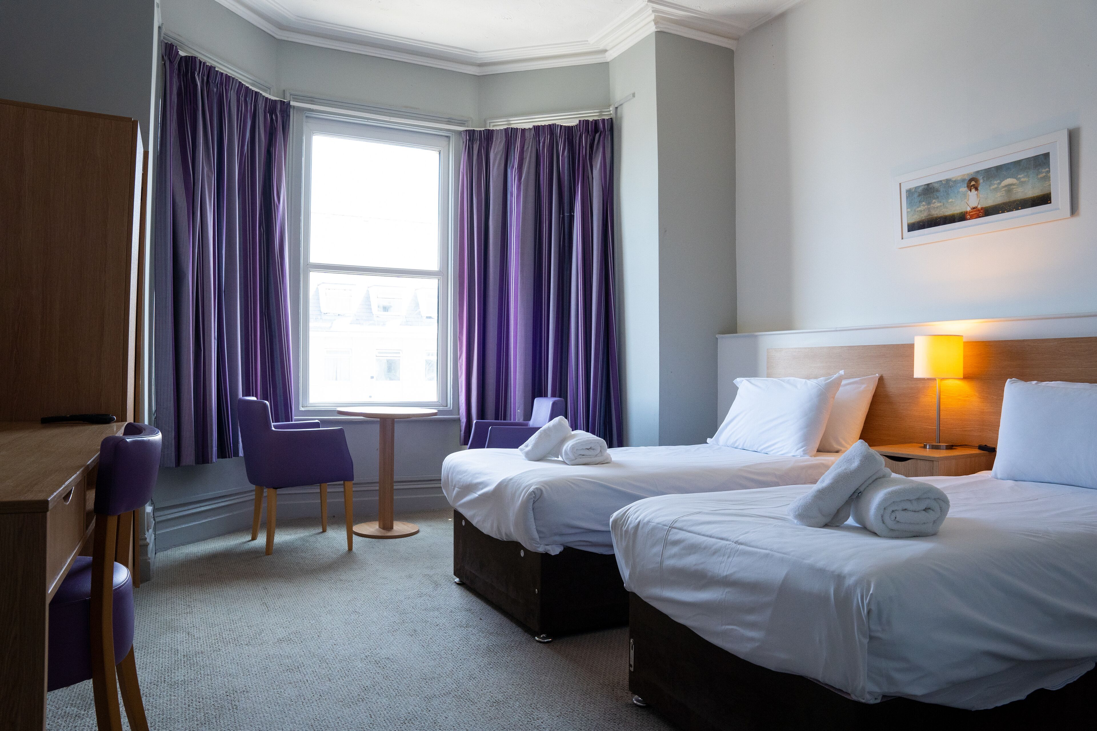 A hotel room with two beds, a desk, a chair, and a window with purple curtains.