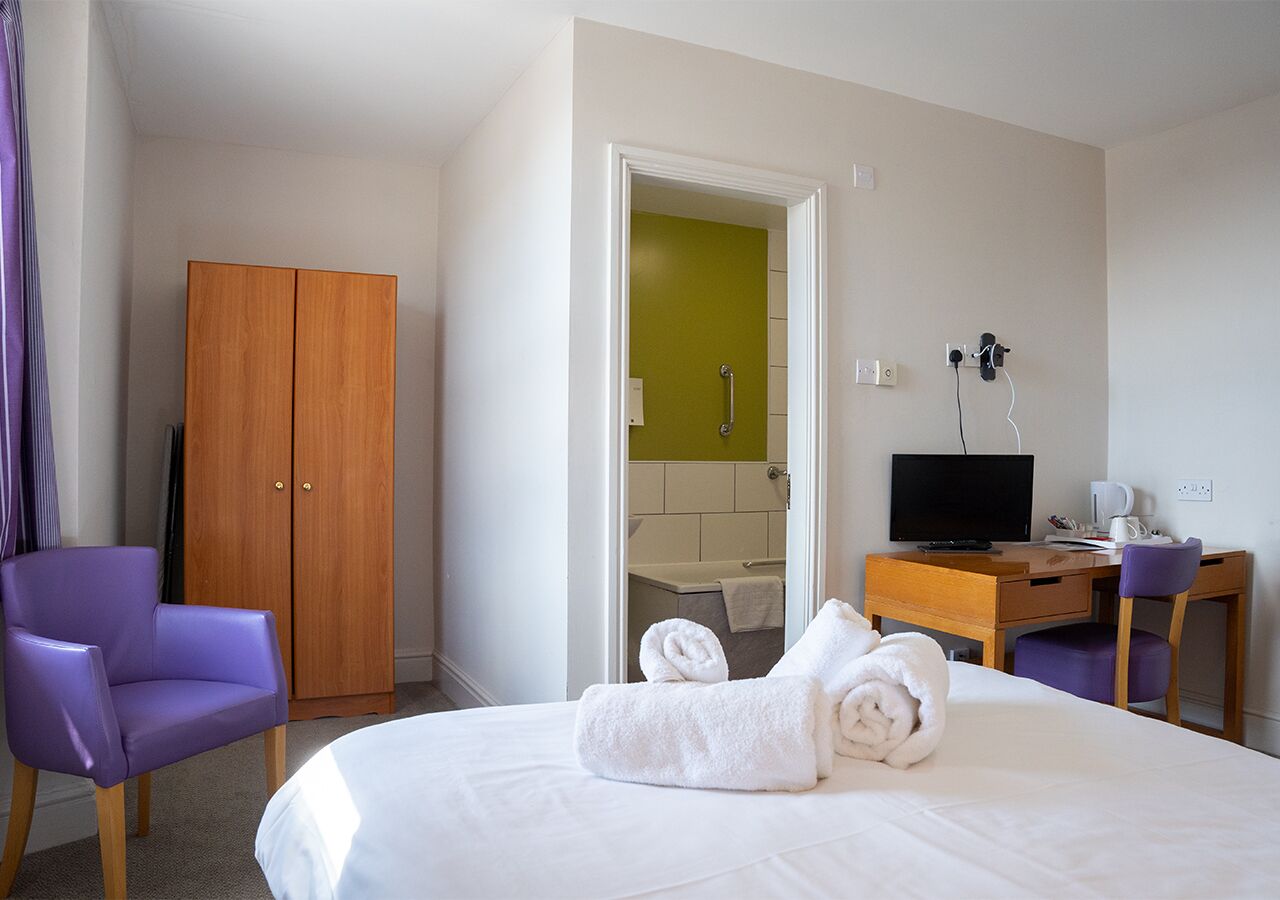 A hotel room with a bed, a desk with a computer, a chair, a wardrobe, and a bathroom visible through an open door.