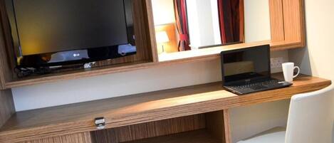 A wooden desk with a laptop and a TV, a white chair, and a cup.