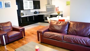 A living room with a brown leather sofa, a smaller armchair, and a wooden coffee table. A kitchen is visible in the background with black cabinets and stainless steel appliances.