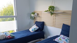 A bedroom with two beds, each with a blue bedspread and colorful pillows. There is a window providing natural light, and a wooden slat headboard with plants on shelves above the beds. - TY SAM (3CH) - RECENT MODERN HOUSE CLOSE TO BEACHES AND AMENITIES (Trébeurden)
