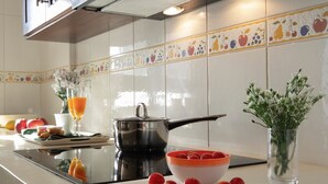Uma cozinha com fogão, panela, tigela de tomates, copo de suco de laranja e um vaso com flores.