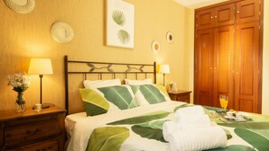 Uma cama bem arrumada com colcha verde e branca listrada, duas luminárias de cabeceira, uma mesinha de cabeceira de madeira com um vaso de flores e um guarda-roupa de madeira.