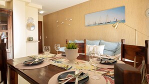Uma área de refeições com mesa de madeira posta para quatro pessoas, um sofá e um quadro de veleiros na parede.