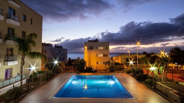 Uma piscina retangular com uma escada de metal, rodeada por um passeio de azulejos. Palmeiras e um edifício com varandas aparecem ao fundo.