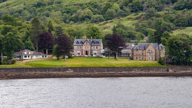 A large, historic building with multiple wings, situated on a lush green hillside beside a body of water. - Claymore Hotel (Arrochar)