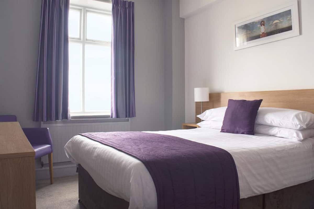 A hotel room with a bed, a chair, a desk, and a window with curtains.