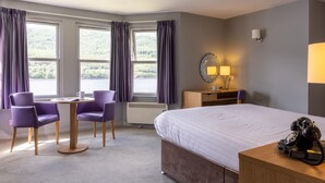 A hotel room with a bed, a desk with a lamp, a telephone, and a view of the outdoors through large windows. - Claymore Hotel (Arrochar)
