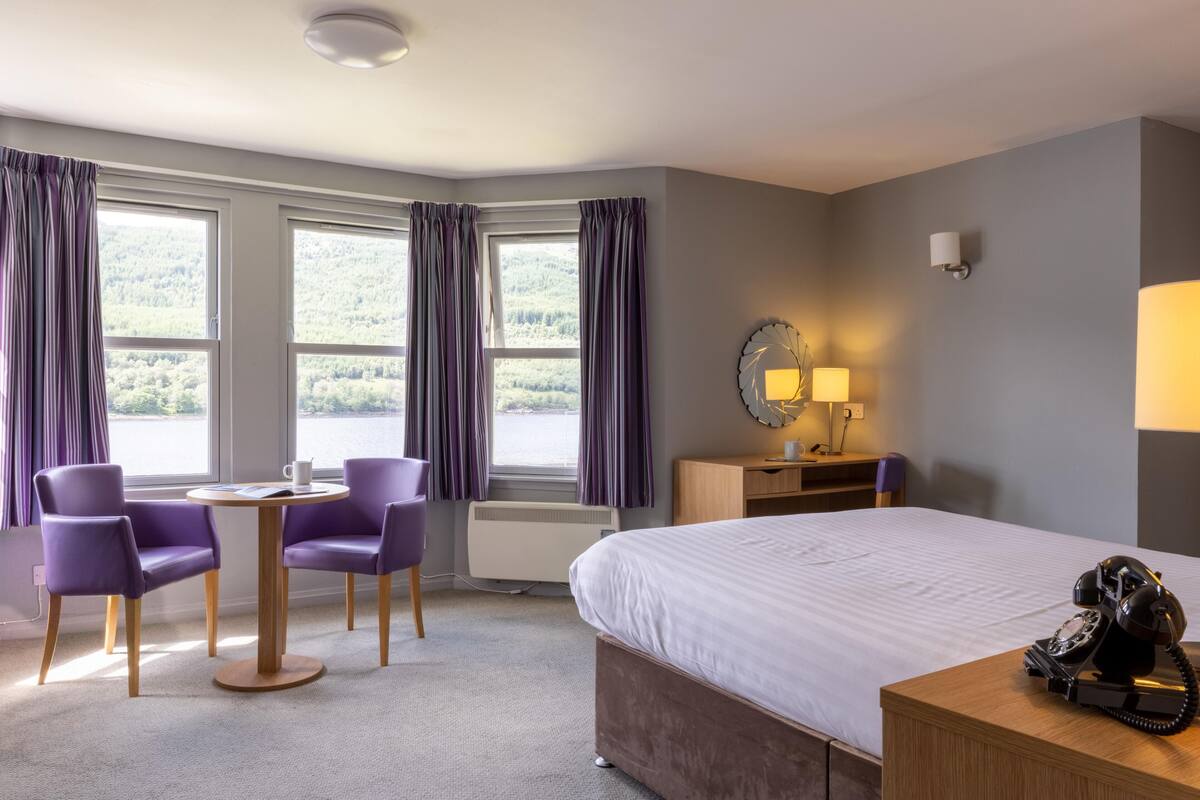 A hotel room with a bed, a desk with a lamp, a telephone, and a view of the outdoors through large windows.