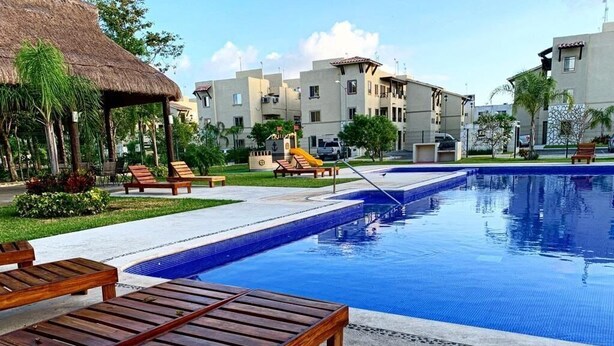 Ein Swimmingpool mit Liegestühlen, einer Struktur mit Reetdach und modernen Gebäuden im Hintergrund.