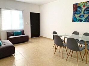 A modern living room with a glass dining table, grey sofas, and a colorful abstract painting on the wall.