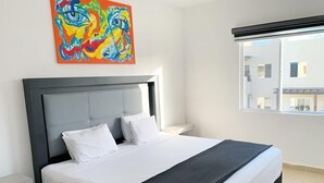 Ein Hotelzimmer mit einem großen Bett, einem Kopfteil, einem Nachttisch, einem Fenster mit Jalousien und einem farbenfrohen abstrakten Gemälde an der Wand.