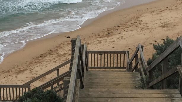 Eine Holztreppe führt zu einem Sandstrand, im Hintergrund sind Meereswellen zu sehen.