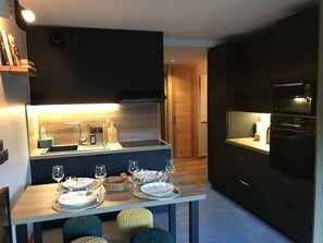 Une cuisine moderne avec des meubles de couleur sombre, un plan de travail en bois et un coin repas aménagé pour quatre personnes, avec des verres à vin et des assiettes.