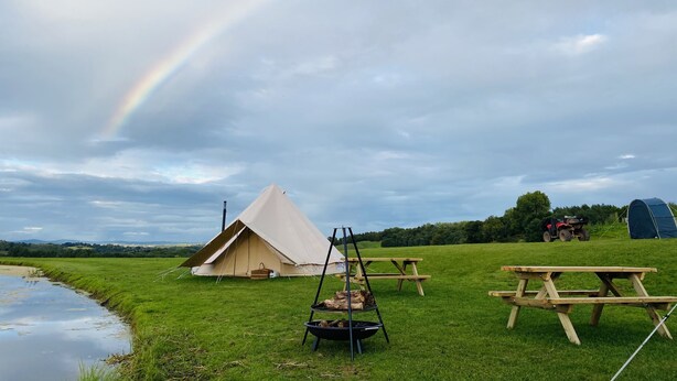 Une tente, une table de pique-nique et un foyer, installés dans un champ verdoyant, avec un arc-en-ciel dans le ciel.