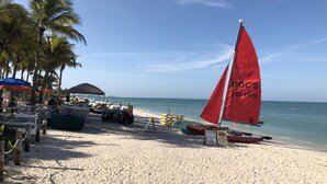 Een strand met een rode zeilboot, palmbomen en strandparasols.