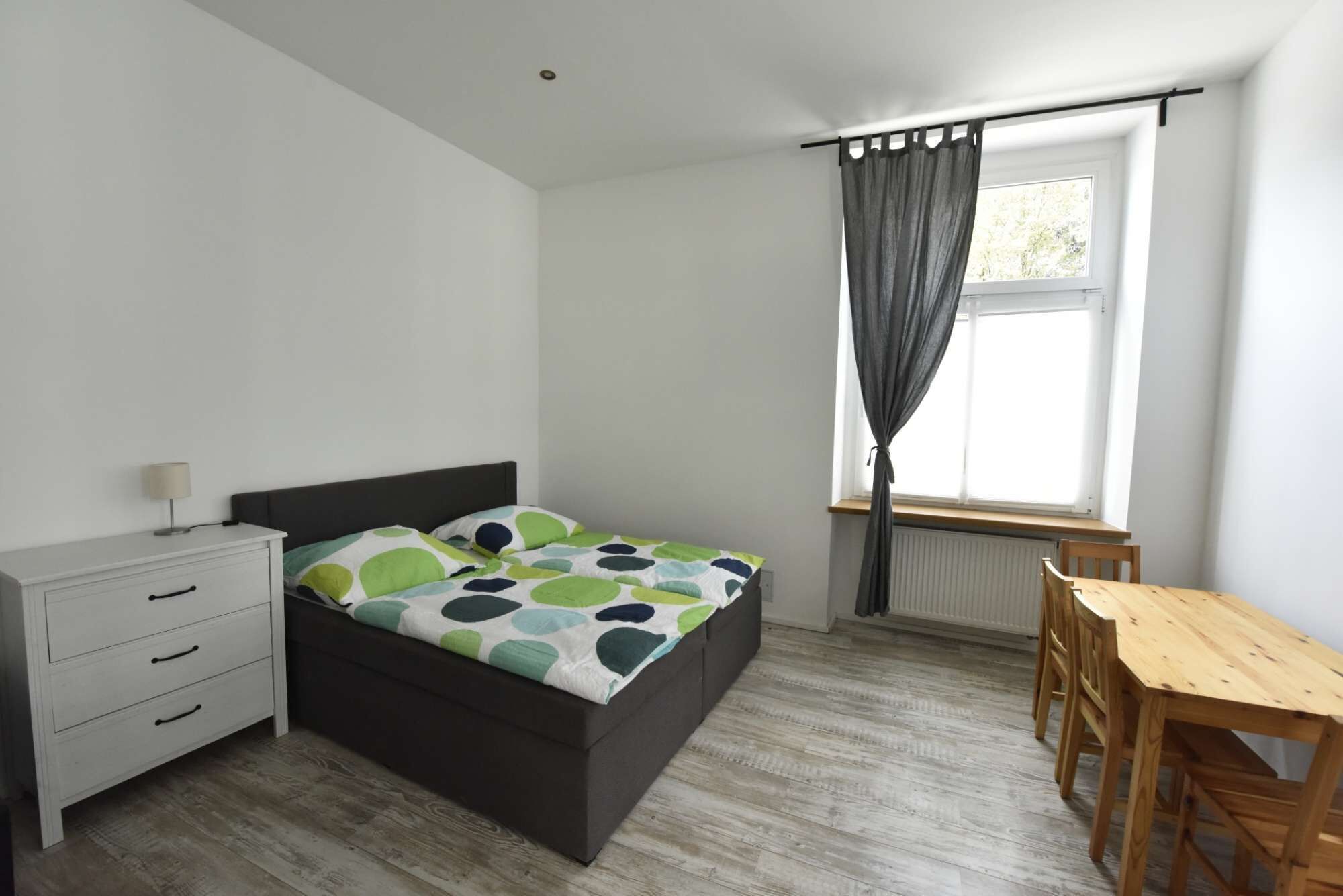A bedroom with a bed, a wooden table, a chair, a nightstand, and a window with curtains.