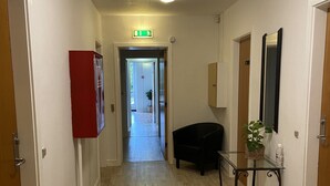 Un pasillo de hotel con suelo de madera, un sillón negro, una mesa de vidrio con una planta y un aparador rojo.