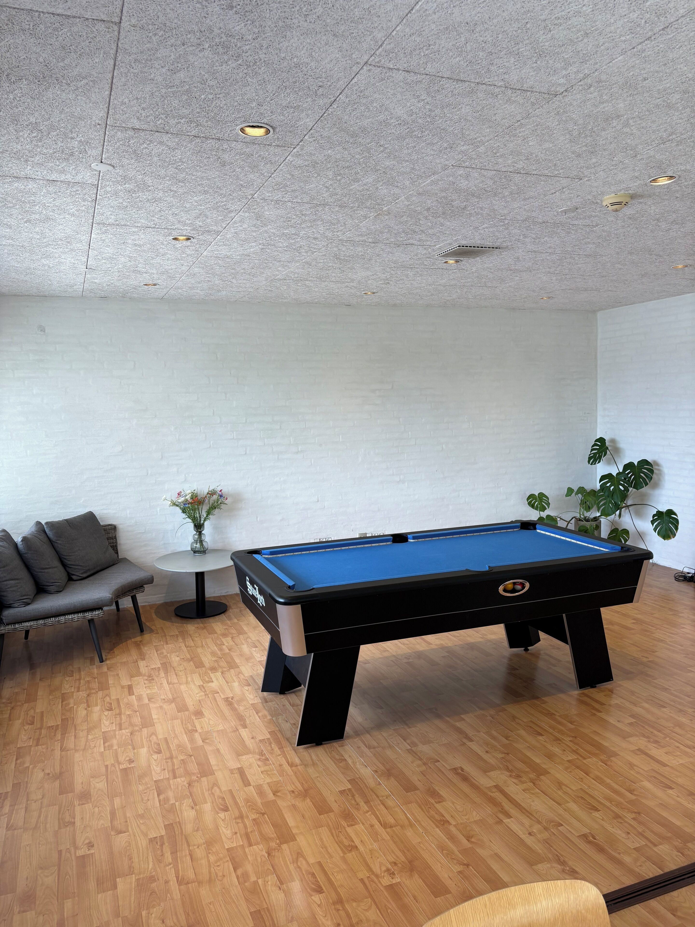 A pool table in a room with a white brick wall, a round table with a vase of flowers, and a sofa.