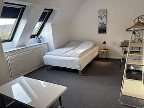 A bedroom with a bed, a table, a radiator, a window, and a wall-mounted shelf.