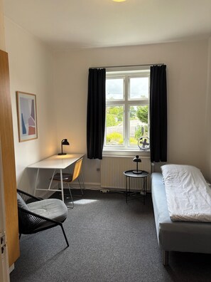 A small, neatly arranged room with a bed, a desk, a chair, and a window with curtains.