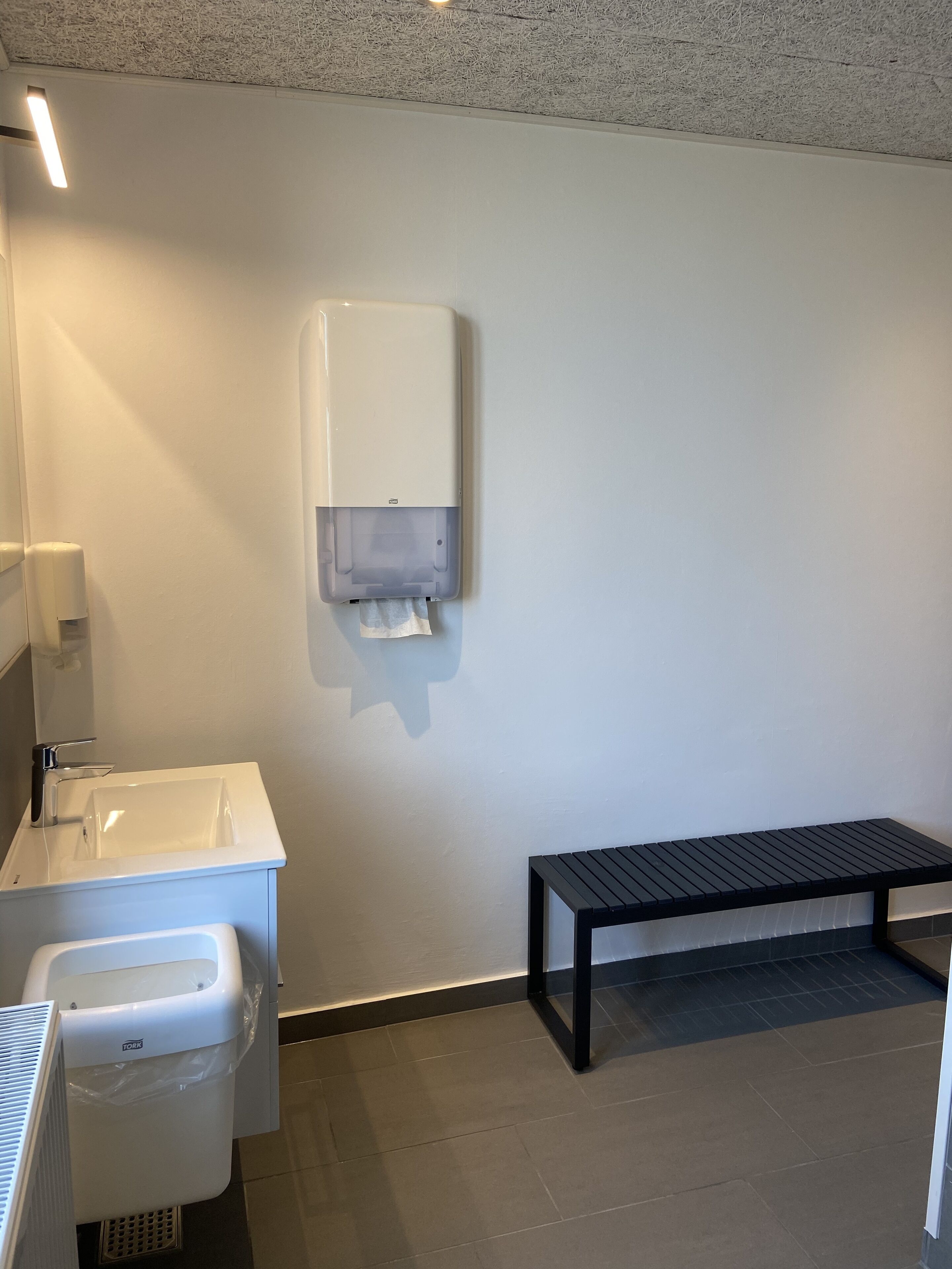 Un baño con lavamanos, secador de manos, cubo de basura y banco.