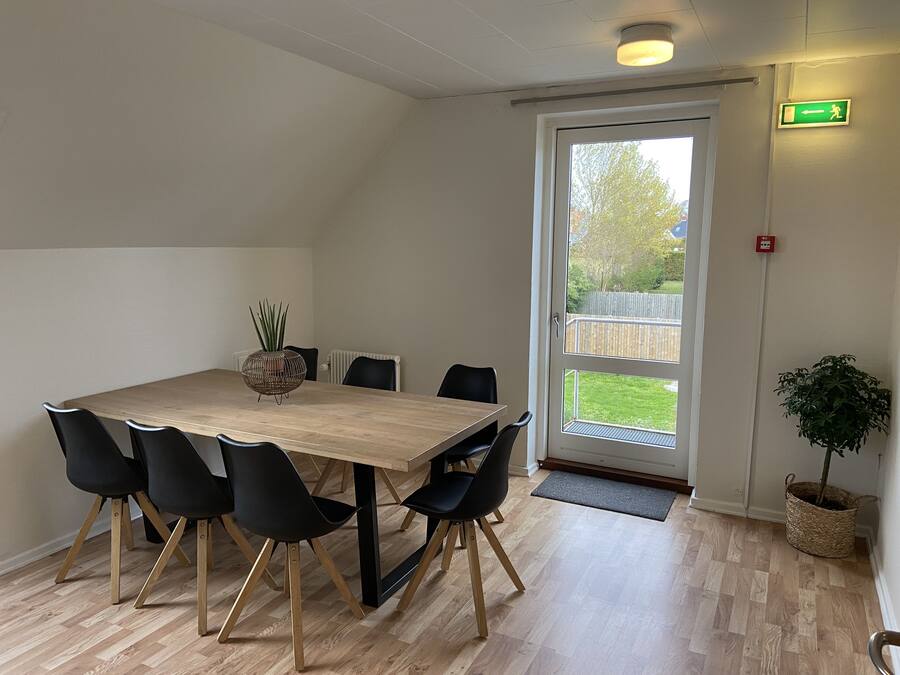 Un coin repas avec une table en bois et des chaises noires, une plante en pot et une vue sur l’extérieur grâce à une porte vitrée.