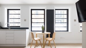 Eine moderne Küche mit weißen Schränken, einer schwarzen Arbeitsplatte und einem runden Tisch mit zwei Stühlen. Zwei Fenster mit schwarzen Vorhängen und ein Blumentopf auf der Fensterbank.