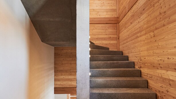 Un escalier en bois avec un palier en béton, menant à une pièce aux murs blancs et au plafond en bois.
