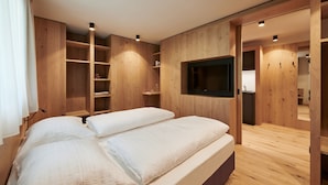 Une chambre moderne avec un plancher en bois, un lit recouvert de draps blancs, des étagères intégrées et un téléviseur fixé au mur.