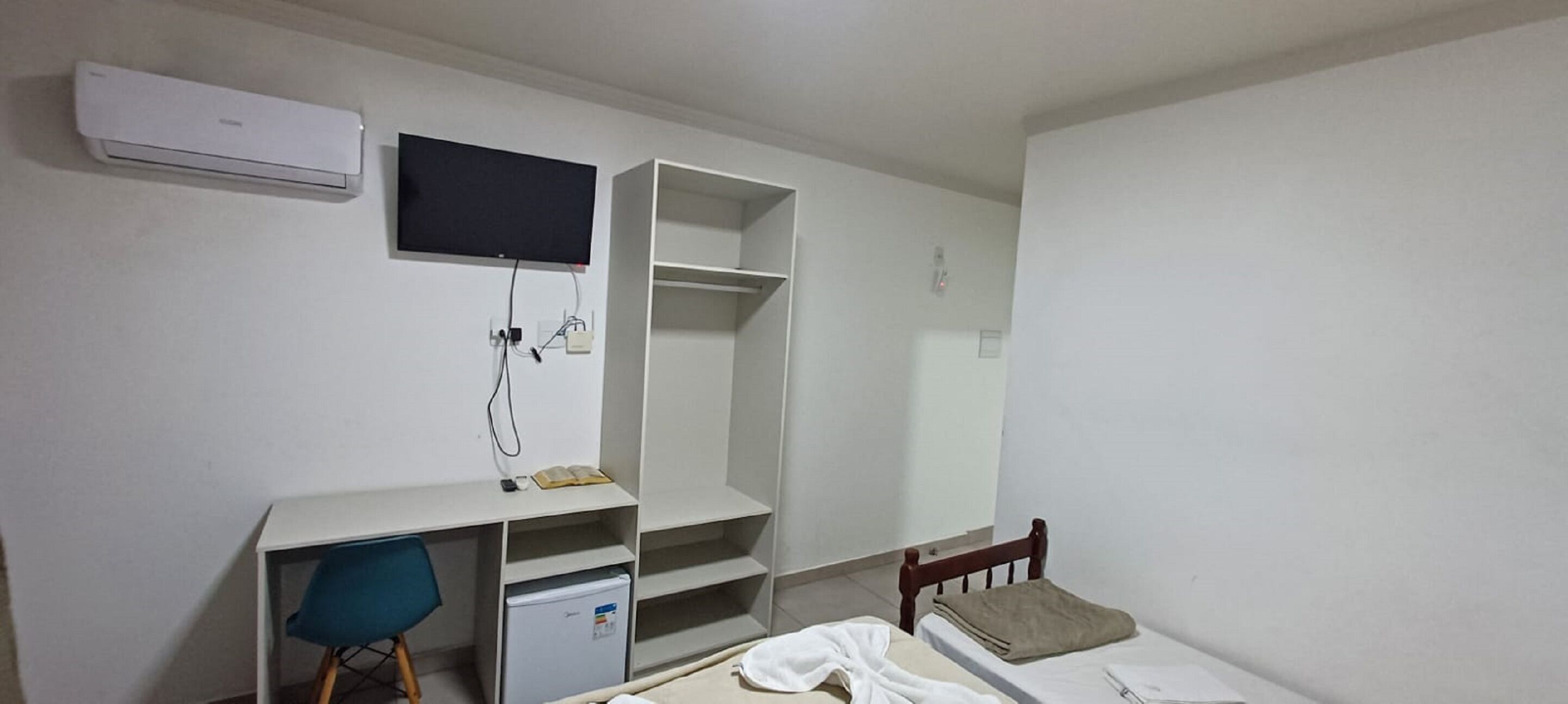 Una habitación pequeña y poco amoblada con escritorio, silla, cama y televisor fijado a la pared.