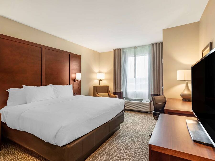 A hotel room with a large bed, a desk with a flat-screen TV, a chair, a lamp, and a window with curtains.