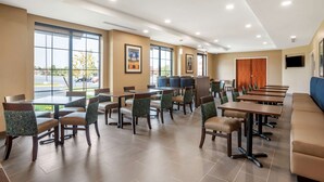 Ein Hotelrestaurant mit mehreren Tischen, Stühlen, großen Fenstern und einem Fernsehbildschirm.