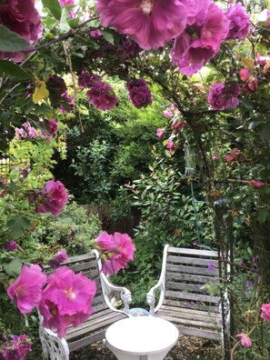 Un jardín con un banco de madera y una mesa, rodeado de abundante vegetación y plantas en flor.
