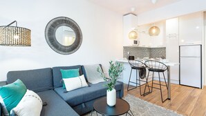 Un salon moderne avec un canapé gris, un miroir rond et une petite table sur laquelle est posée une plante. On aperçoit à l’arrière-plan un coin cuisine avec un tabouret de bar et un réfrigérateur.