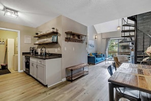 Una cocina moderna con estantes de madera, un comedor con mesa de madera y una escalera que sube a un altillo.