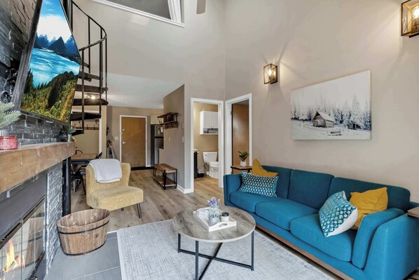 Un salon moderne avec un canapé bleu, une cheminée, un escalier et un tableau représentant un paysage enneigé.
