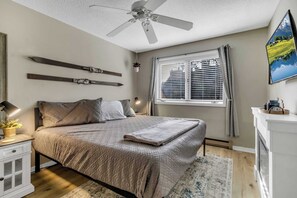 Un dormitorio con cama, ventilador de techo, ventana con persianas, mesita de noche con lámpara y un cuadro en la pared.