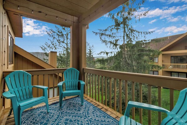 Um espaço ao ar livre coberto com duas cadeiras Adirondack turquesa, um tapete estampado e vista para árvores e edifícios ao redor.