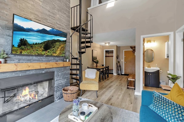 A modern living room with a fireplace, a large TV, a staircase, and a dining area. - SnowPine Condo, Sleeps 6, Patio, A/C (Ellicottville)