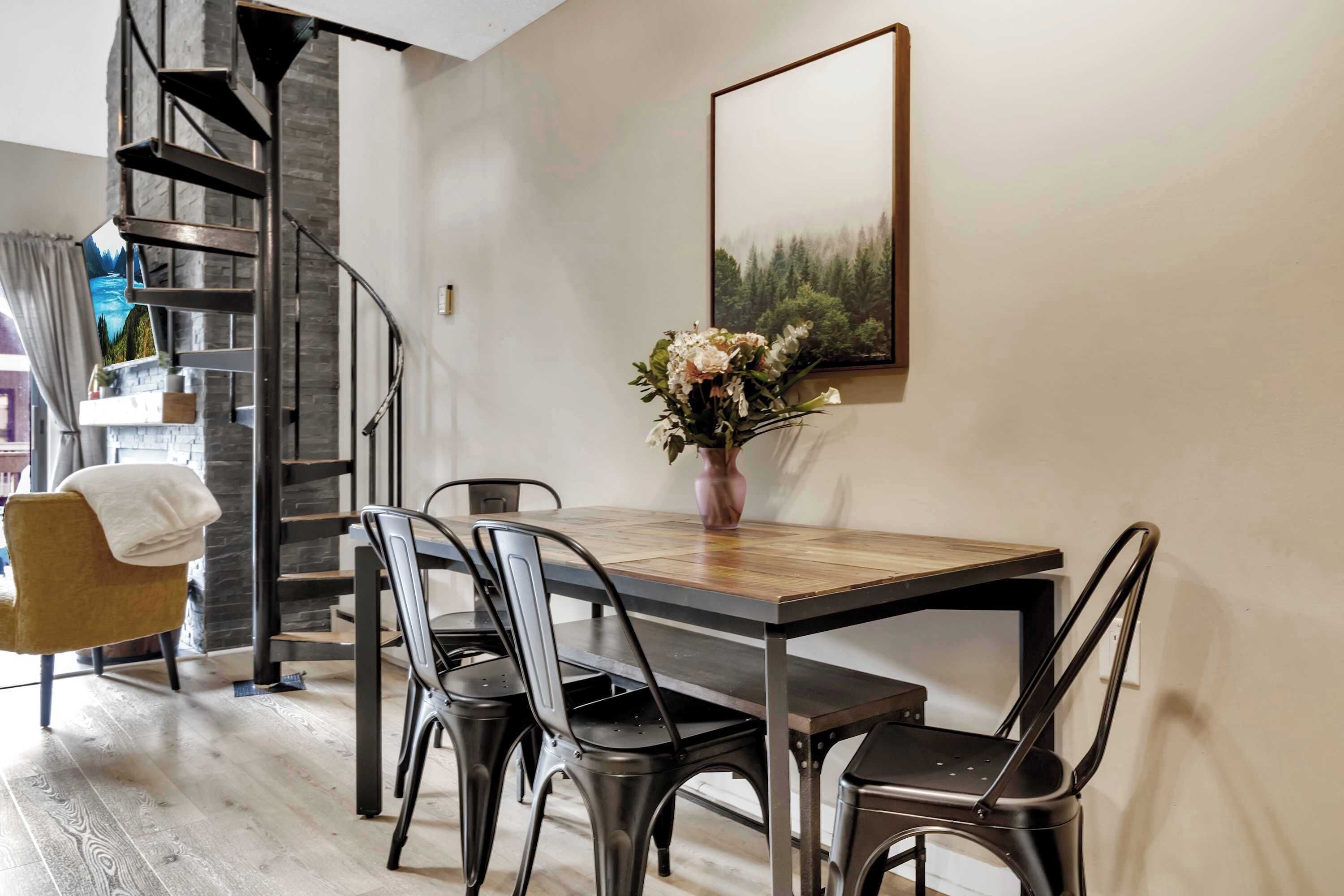 Un coin repas avec une table en bois, des chaises en métal, un vase avec des fleurs, un escalier et un tableau encadré au mur.