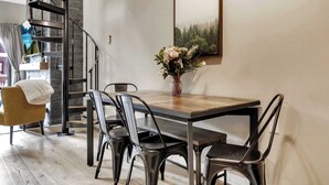 Uma área de refeições com uma mesa de madeira, cadeiras de metal, um vaso com flores, uma escada e um quadro na parede.