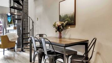 Un coin repas avec une table en bois, des chaises en métal, un vase avec des fleurs, un escalier et un tableau encadré au mur.