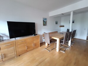 A modern living room with a flat-screen TV, wooden cabinets, a dining table with chairs, and a kitchen area.