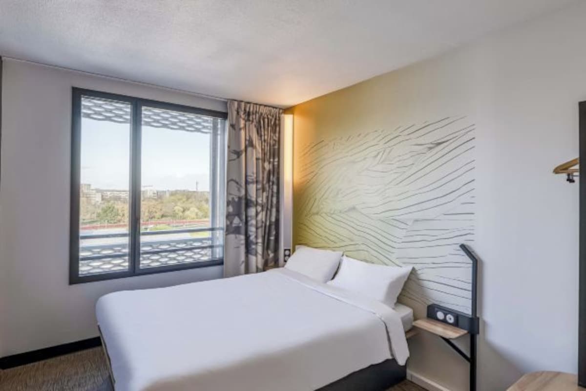A hotel room with a large bed, a window with a view, a headboard with a textured design, and a bedside table with a lamp.