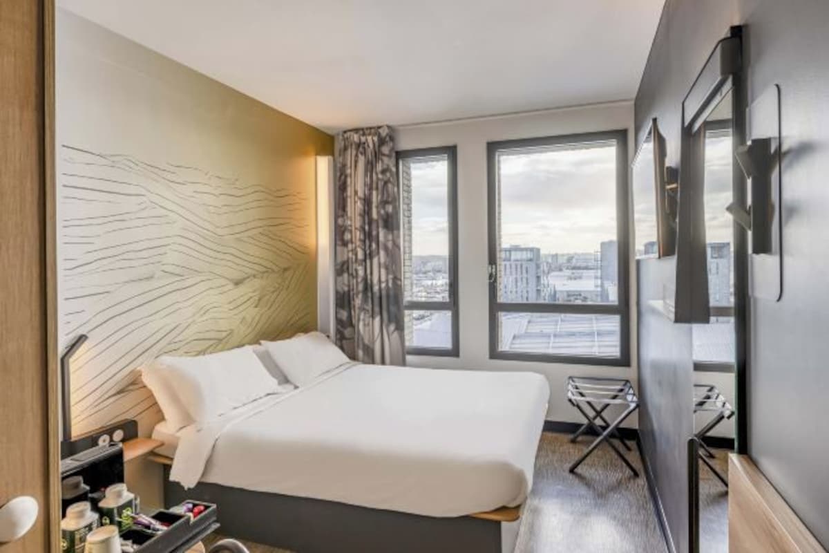 A modern hotel room with a large bed, a desk, and a city view through the windows.