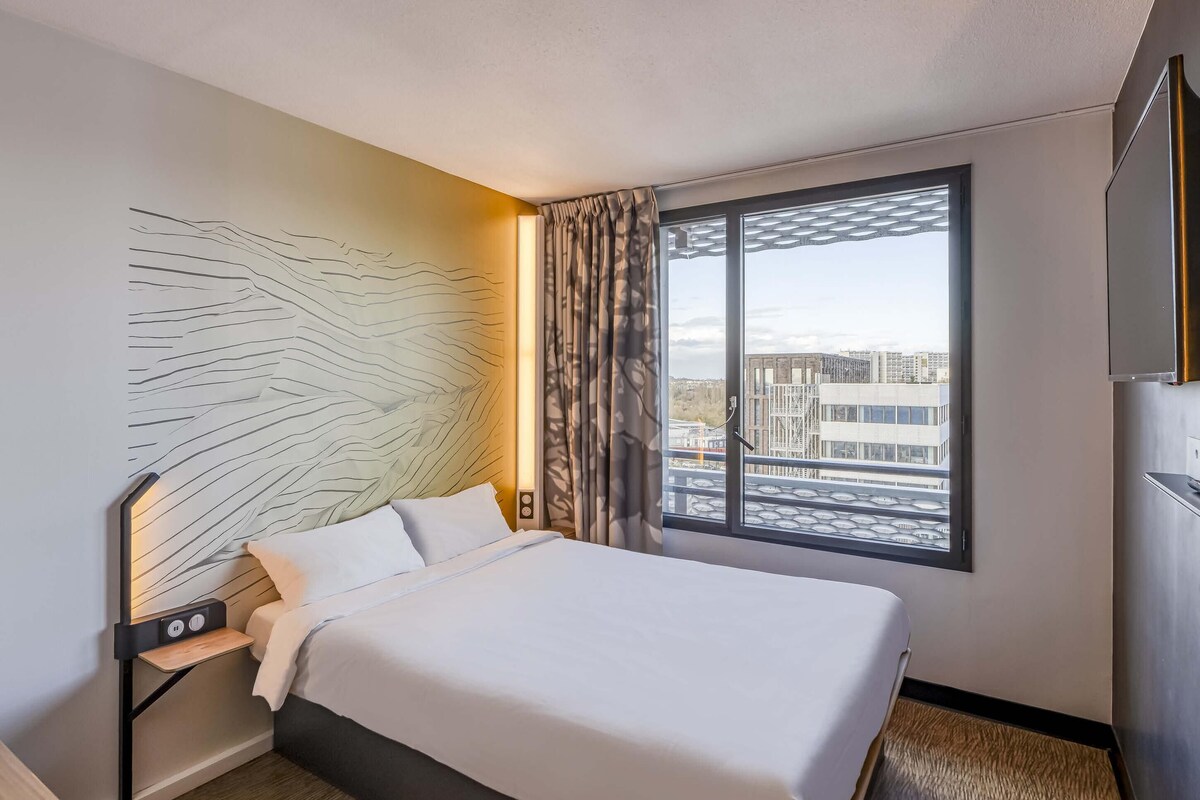 Ein Hotelzimmer mit einem Bett, einem Nachttisch mit Lampe, einem großen Fenster mit Blick und einem Wandbild.